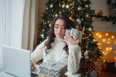 Açık renk örgü kazaklı mutlu, siyah saçlı kıvırcık kadın Noel ağacının yanında hediyeler hazırlıyor. Şık bir model kış tatilinde online alışveriş yapıyor. Yeni yıl. Konforlu ev.