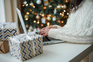 Açık renk örgü kazaklı mutlu, siyah saçlı kıvırcık kadın Noel ağacının yanında hediyeler hazırlıyor. Şık bir model kış tatilinde online alışveriş yapıyor. Yeni yıl. Konforlu ev.