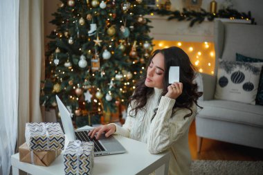 Açık renk örgü kazaklı mutlu, siyah saçlı kıvırcık kadın Noel ağacının yanında hediyeler hazırlıyor. Şık bir model kış tatilinde online alışveriş yapıyor. Yeni yıl. Konforlu ev.