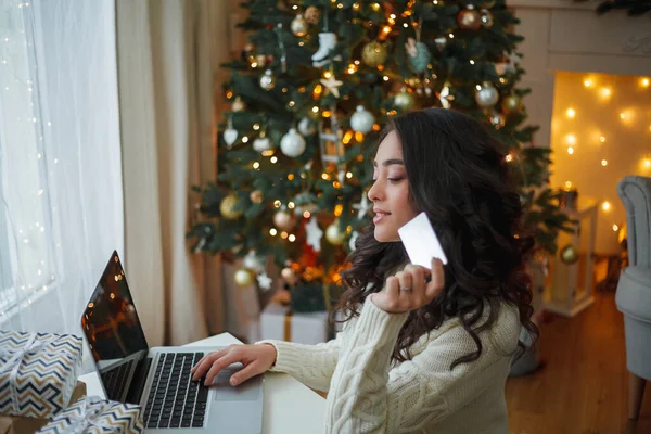 Feliz joven morena rizada mujer en un suéter de punto ligero prepara regalos cerca del árbol de Navidad. Elegante modelo hace compras en línea durante las vacaciones de invierno. Año Nuevo. — Foto de Stock