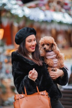 Sahte kürklü ve bereli mutlu, genç, siyah saçlı bir kadın bir Avrupa şehrinin caddesinde köpek kayısı küçük kanişiyle alışveriş yapıyor. Kış tatili Noel.