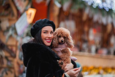 Sahte kürklü ve bereli mutlu, genç, siyah saçlı bir kadın bir Avrupa şehrinin caddesinde köpek kayısı küçük kanişiyle alışveriş yapıyor. Kış tatili Noel.