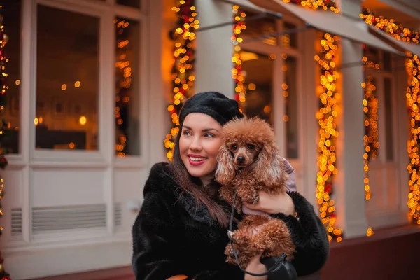 Feliz joven mujer morena emocional en abrigo de piel sintética y boina de compras con su perro de albaricoque pequeño caniche en la calle de una ciudad europea. Vacaciones de invierno. Navidad. — Foto de Stock