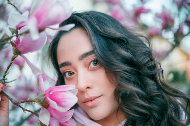 Spring portrait of young dark haired curly woman near blooming magnolia. Emotional model near tree with pink flowers. Warm season. Holidays.