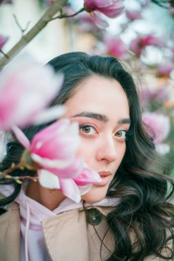 Spring portrait of young dark haired curly woman near blooming magnolia. Emotional model near tree with pink flowers. Warm season. Holidays.