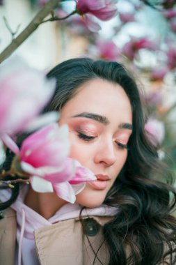 Spring portrait of young dark haired curly woman near blooming magnolia. Emotional model near tree with pink flowers. Warm season. Holidays.