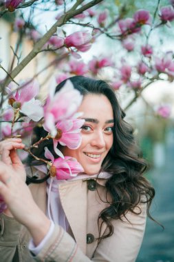 Spring portrait of young dark haired curly woman near blooming magnolia. Emotional model near tree with pink flowers. Warm season. Holidays.