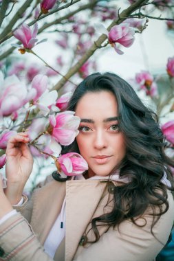 Spring portrait of young dark haired curly woman near blooming magnolia. Emotional model near tree with pink flowers. Warm season. Holidays.