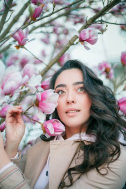 Spring portrait of young dark haired curly woman near blooming magnolia. Emotional model near tree with pink flowers. Warm season. Holidays.