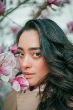 Spring portrait of young dark haired curly woman near blooming magnolia. Emotional model near tree with pink flowers. Warm season. Holidays.