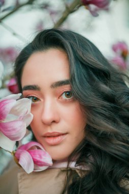 Spring portrait of young dark haired curly woman near blooming magnolia. Emotional model near tree with pink flowers. Warm season. Holidays.