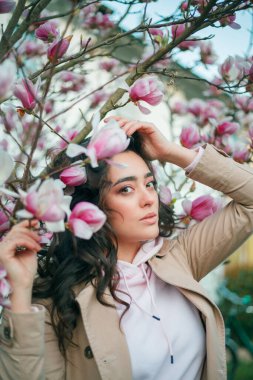Spring portrait of young dark haired curly woman near blooming magnolia. Emotional model near tree with pink flowers. Warm season.