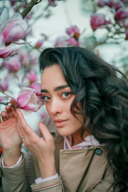 Spring portrait of young dark haired curly woman near blooming magnolia. Emotional model near tree with pink flowers. Warm season.