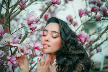 Spring portrait of young dark haired curly woman near blooming magnolia. Emotional model near tree with pink flowers. Warm season.