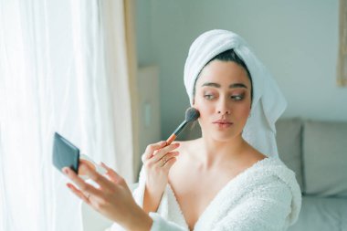 Young beautiful woman in terry white dressing gown makes herself beautiful makeup at home. Model holds makeup brush and cosmetics in her hands. Cozy home.