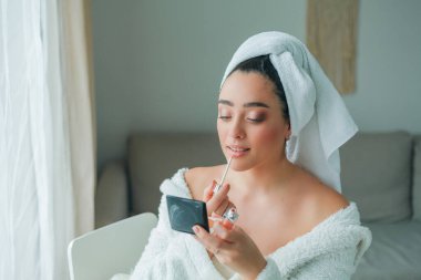 Young beautiful woman in terry white dressing gown makes herself beautiful makeup at home. Model holds makeup brush and cosmetics in her hands. Cozy home.