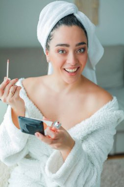 Young beautiful woman in terry white dressing gown makes herself beautiful makeup at home. Model holds makeup brush and cosmetics in her hands. Cozy home.