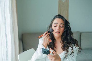 Young beautiful woman in terry white dressing gown makes herself beautiful makeup at home. Model holds makeup brush and cosmetics in her hands. Cozy home.