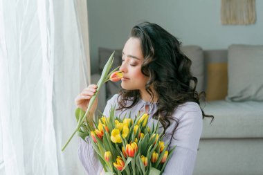 İlkbahar portresi, güzel, koyu renk kıvırcık saçlı, leylak elbiseli bir kadın. Evde büyük bir buket lale var. Duygusal model elinde çiçekleri tutuyor. Kadınlar Günü. Hediye. Anneler Günü.