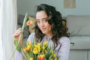 İlkbahar portresi, güzel, koyu renk kıvırcık saçlı, leylak elbiseli bir kadın. Evde büyük bir buket lale var. Duygusal model elinde çiçekleri tutuyor. Kadınlar Günü. Hediye. Anneler Günü.