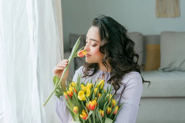 İlkbahar portresi, güzel, koyu renk kıvırcık saçlı, leylak elbiseli bir kadın. Evde büyük bir buket lale var. Duygusal model elinde çiçekleri tutuyor. Kadınlar Günü. Hediye. Anneler Günü.