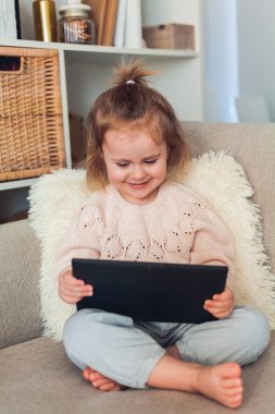 Okul online. 3 yaşındaki sevimli küçük kız rahat bir evde kanepede oturuyor ve bir video görüşmesinde konuşuyor. Elinde tablet tutan duygusal bir çocuğun portresi. Ev.