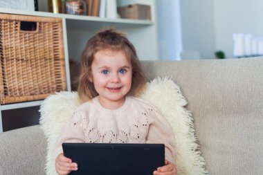 Okul online. 3 yaşındaki sevimli küçük kız rahat bir evde kanepede oturuyor ve bir video görüşmesinde konuşuyor. Elinde tablet tutan duygusal bir çocuğun portresi. Ev.
