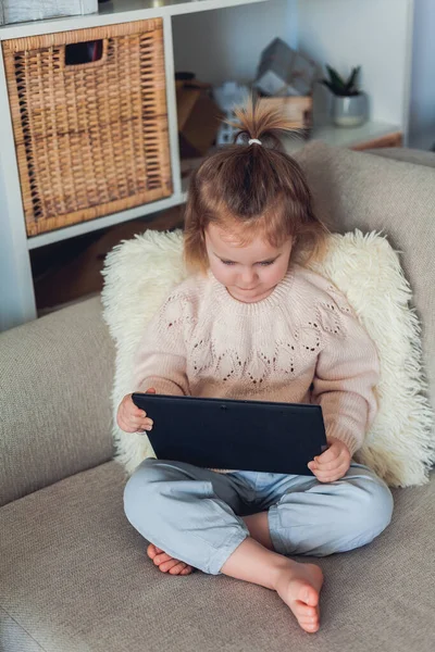 Okul online. 3 yaşındaki sevimli küçük kız rahat bir evde kanepede oturuyor ve bir video görüşmesinde konuşuyor. Elinde tablet tutan duygusal bir çocuğun portresi. Ev.