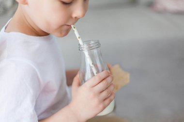 Açık renk elbiseli sevimli, güzel saçlı çocuk cam şişeden süt içiyor ve evde bir somun ekmek yiyor. Eko ürünler. Sağlıklı yiyecek ve içecekler. Süt.