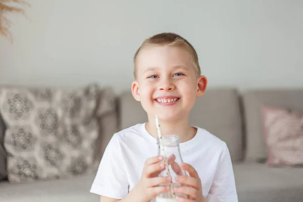 明るい服を着た愛らしい幸せなフェアヘアの少年は ガラス瓶から牛乳を飲み 自宅でパンのローフを食べる エコ製品 健康的な食べ物や飲み物 ミルク — ストック写真