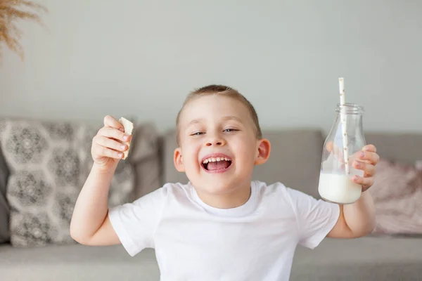 明るい服を着た愛らしい幸せなフェアヘアの少年は ガラス瓶から牛乳を飲み 自宅でパンのローフを食べる エコ製品 健康的な食べ物や飲み物 ミルク — ストック写真