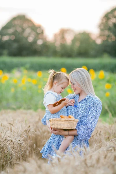 Adorable mother and daughter in romantic dresses eat fresh pastries in a wheat field. Summer family portrait. Eco. — Stock Photo, Image