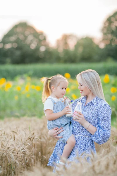 Adorable mother and daughter in romantic dresses drink milk from glass bottles with paper tubes in a wheat field. Summer family portrait. Eco. — Stock Photo, Image