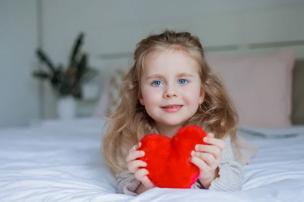 青い目の3歳の愛らしい少女は 彼女の手の中に赤いぬいぐるみの心を持っています バレンタインデー 感情的な子供だ ホーム — ストック写真