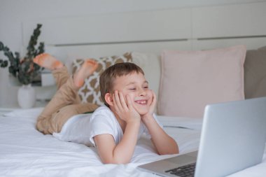 Sevimli okul çocuğu evde yatakta yatarken internet kullanıyor. Online okul. 7 yaşında tatlı bir çocuk dizüstü bilgisayarda oyun oynuyor. Konforlu ev.