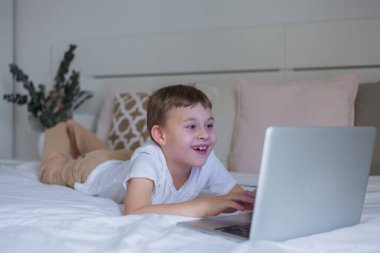 Sevimli okul çocuğu evde yatakta yatarken internet kullanıyor. Online okul. 7 yaşında tatlı bir çocuk dizüstü bilgisayarda oyun oynuyor. Konforlu ev.