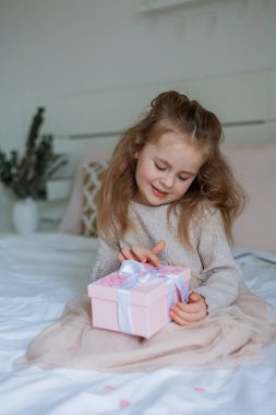 Şirin mavi gözlü kız elinde pembe bir fiyonk olan bir hediye kutusu tutuyor. Mutlu çocuk annesine bir hediye verir. Doğum günü. Anneler Günü. Ev.