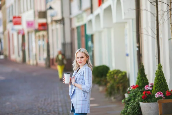 Hermosa joven rubia bebe café de una taza de papel y pasea por la ciudad. Retrato de una modelo joven y elegante en ropa casual al aire libre. Primavera. — Foto de Stock
