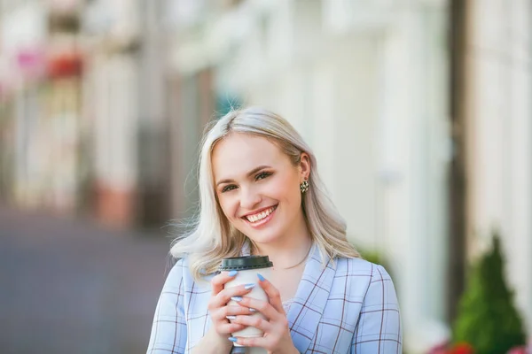 Vacker ung blond kvinna dricker kaffe från en pappersmugg och går runt i staden. Porträtt av en stilren ung modell i avslappnade kläder utomhus. Våren. — Stockfoto