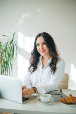 Genç iş kadını bilgisayarda çalışıyor. Beyaz gömlekli, güzel, koyu renk saçlı bir kadın internette, parlak bir ofiste çalışıyor. Kahve içip kruvasan yemek. Ofis.