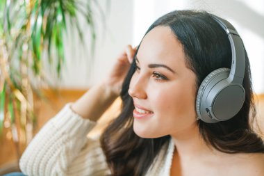 Genç, siyah saçlı, örgü hırkalı, güzel kıvırcık kadın rahat bir evde koltukta otururken kulaklıkla müzik dinliyor. Dinlen..