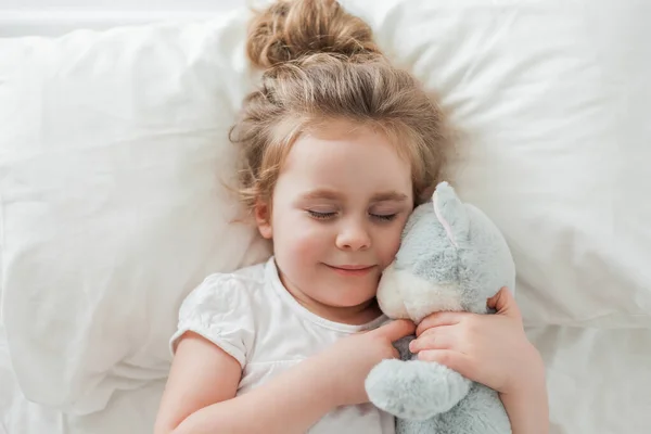 Niña linda con el pelo rizado duerme en la cama en una cama blanca con su juguete de peluche. Un sueño saludable. El niño está descansando.. — Foto de Stock