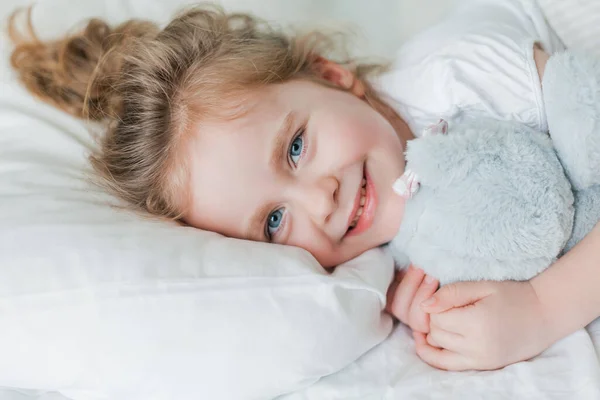 Piccola ragazza carina con i capelli ricci dorme a letto su un letto bianco con il suo peluche giocattolo. Sonno sano. Il bambino sta riposando. — Foto Stock
