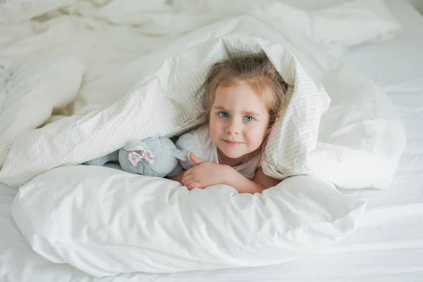 Piccola ragazza riccia dagli occhi azzurri carina gioca a letto con il suo peluche giocattolo. Il bambino è sdraiato nel letto sotto le coperte. Emozioni. Sonno sano. — Foto Stock
