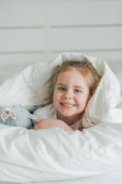 Piccola ragazza riccia dagli occhi azzurri carina gioca a letto con il suo peluche giocattolo. Il bambino è sdraiato nel letto sotto le coperte. Emozioni. Sonno sano. — Foto Stock