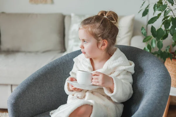 Pequeña chica de pelo rizado en una bata de luz suave bebe té y se relaja en casa sentado en una silla cómoda. El bebé imita a su madre. Hermoso acogedor interior acogedor. — Foto de Stock