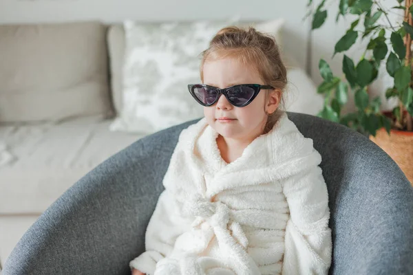 Piccola ragazza carina di 3 anni in morbido accappatoio nei suoi occhiali madri si siede in una poltrona a casa e gioca in un salone di bellezza. Riposo. — Foto Stock