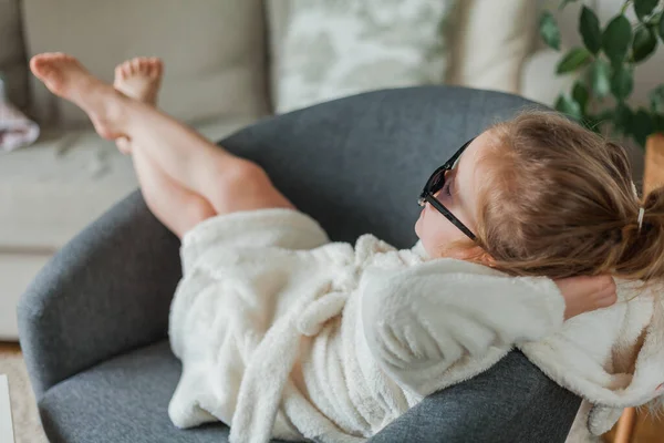Niña linda de 3 años de edad en albornoz de felpa en sus gafas madres se sienta en un sillón en casa y juega en un salón de belleza. Descanso. — Foto de Stock