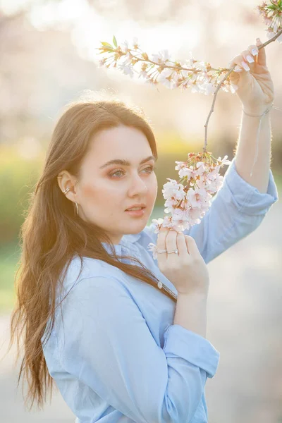 Bela jovem de 19 anos em roupas casuais leves em tons pastel perto de árvores de sakura floridas. Retrato de primavera de modelo com olhos azuis. — Fotografia de Stock