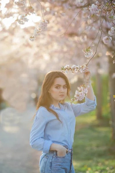 Красива молода жінка 19-річного віку в легкому повсякденному одязі в пастельних відтінках біля квітучих сакури. Весняний портрет моделі з блакитними очима . — стокове фото